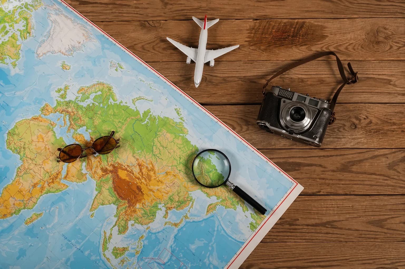 Mapa-múndi aberto sobre mesa de madeira com miniatura de avião, câmera fotográfica e óculos de sol
